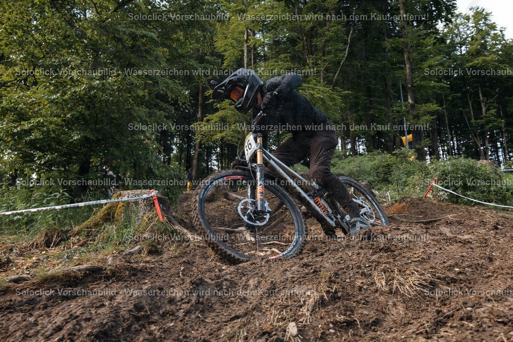 Mountainbike-Action im Wald | Ein Mountainbiker in voller Ausrüstung rast einen staubigen, holprigen Pfad hinunter. Er trägt einen schwarzen Helm mit Visier, eine schwarze Jacke und schwarze Handschuhe. Das Gelände ist uneben und mit Erde bedeckt, und es gibt Bäume und Büsche im Hintergrund. Das Bild vermittelt ein Gefühl von Geschwindigkeit und Abenteuer. - Realisiert mit Pictrs.com