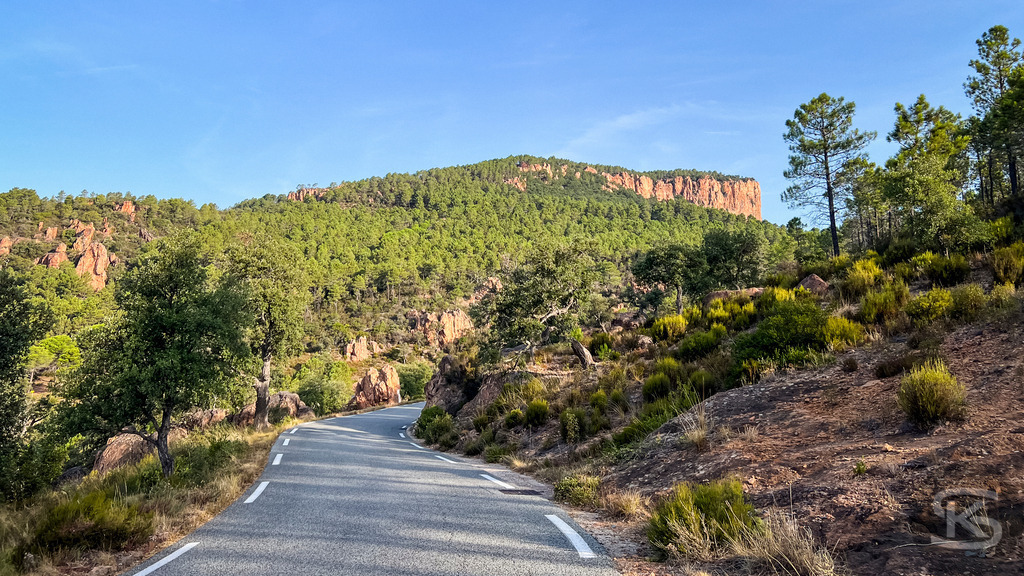Kurvige Landstraße durch das Hinterland von Fréjus und das Esterel-Massiv | Eine schmale Asphaltstraße windet sich durch die mediterrane Hügellandschaft mit grünen Pinien und den charakteristischen roten Felsformationen im Hinterland von Fréjus bei den Gorges de Pennafort. - Realisiert mit Pictrs.com