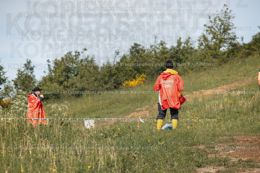 Untersuchung im Freien | Die Aufnahme zeigt zwei Personen, die in einem Feld mit hohem Gras und Büschen stehen. Beide tragen Warnwesten, wobei eine Person einen gelben Regenmantel trägt. Im Vordergrund ist ein schwarzes Absperrband mit gelben Markierungen zu sehen. Die Szene vermittelt einen Eindruck von Aktivität und möglicherweise einer Untersuchung oder Absicherung eines Bereichs. - Realisiert mit Pictrs.com