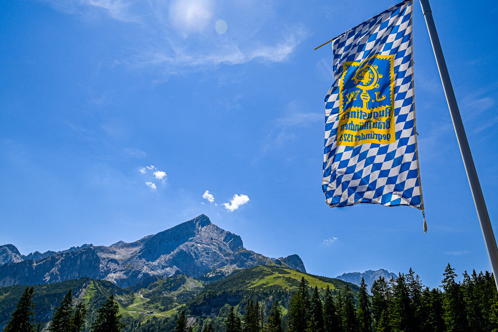 Bayerischer Himmel über der Alpspitze | Das Foto fängt ein echtes Stück Bayern ein. Unter einem strahlend blauen Sommerhimmel mit lockeren Schönwetterwolken ragen die schroffen, grauen Felsen der markanten Alpspitze majestätisch empor. Im Vordergrund weht eine traditionelle weiß-blaue Rautenflagge der Brauerei Augustiner-Bräu München stolz im Wind und unterstreicht die alpine Idylle. Dunkle Tannenwipfel bilden einen natürlichen Rahmen, während grüne Almen am Fuß des Berges das Bild vervollständigen und Sehnsucht nach Bergen wecken. - Realisiert mit Pictrs.com
