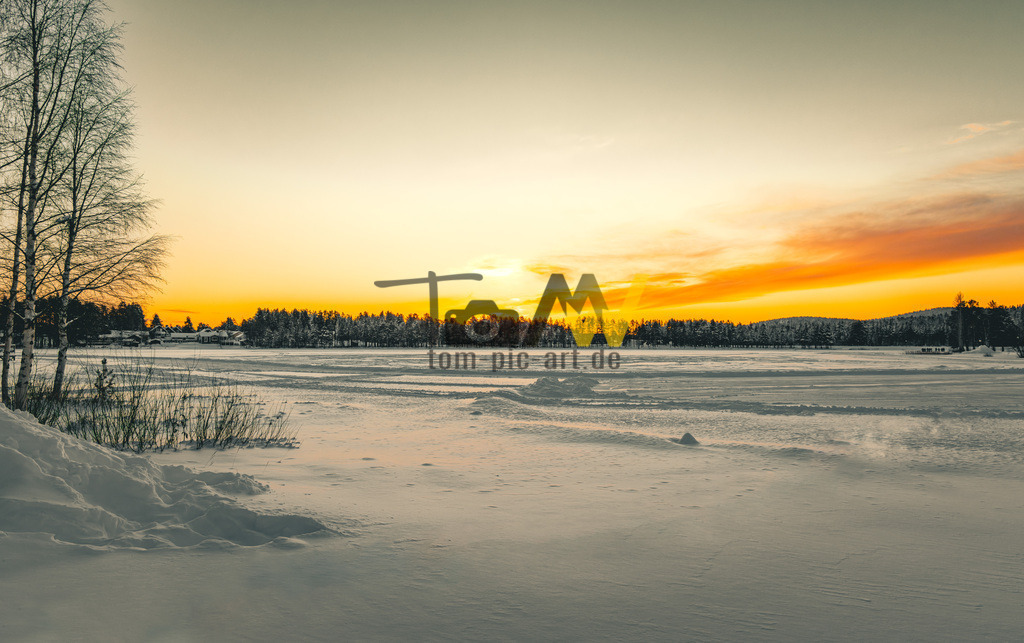 Stille und ruhige Winterszene eine zugefrorenen See's--Eis | Das Bild zeigt einen winterlichen Sonnenaufgang oder Sonnenuntergang in einer schneebedeckten Landschaft, möglicherweise in Lappland. Die Szene zeigt eine weite, gefrorene Fläche, die von Schnee bedeckt ist.Am Horizont sind Bäume und ein leuchtend orangefarbener Himmel zu sehen.Das Bild trägt ein Wasserzeichen mit der Aufschrift "tom-pic-art.de".Die Lichtverhältnisse deuten auf einen Zeitpunkt nahe der Wintersonnenwende hin, an dem die Sonne nur sehr kurz über dem Horizont steht. - Realisiert mit Pictrs.com