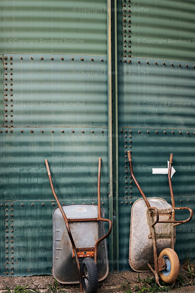 2 Schubkarren | Zwei Schubkarren lehnen an einer Blechwand.  - Realisiert mit Pictrs.com