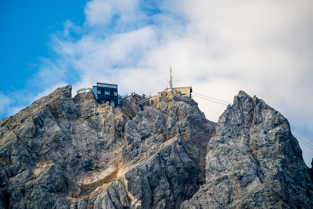 Dem Himmel so nah: Die Zugspitze in all ihrer Erhabenheit | Schroffe Felsformationen und imposante Steinwände ragen kühn in den strahlend blauen Himmel empor. Auf den Gipfeln thronen die markante Bergstation und der Sendemast des Zugspitzmassivs – ein faszinierendes Zusammenspiel aus wilder, unbezwungener Natur und kühner menschlicher Ingenieurskunst. Sanfte Wolkenfelder umspielen die schneidigen Gratlinien und verleihen der mächtigen Kulisse eine beinahe erhabene Ästhetik. Das Foto fängt den Reiz der Extreme, die Sehnsucht nach Höhe und die atemberaubende Erhabenheit der Alpen ein. - Realisiert mit Pictrs.com