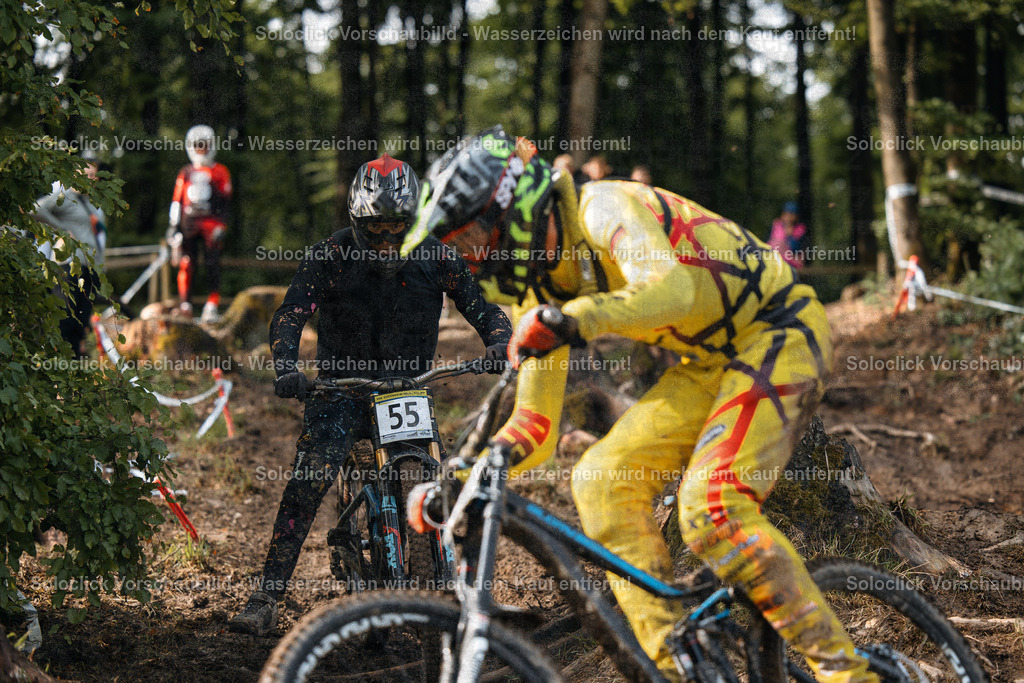 Mountainbike-Abenteuer im Winterwald | Das Bild zeigt zwei Mountainbiker in voller Montur, die einen steilen, verschneiten Waldweg hinabfahren. Beide Fahrer sind mit Schutzhelmen und Schutzkleidung ausgestattet und tragen ihre Startnummern. Der Fahrer links trägt ein schwarzes Trikot und eine schwarze Hose, während der Fahrer rechts ein gelb-grünes Trikot und Hose trägt. Beide Fahrräder sind mit Federgabeln und dicken Reifen ausgestattet, die für das Gelände geeignet sind. Der Hintergrund besteht aus dichtem Wald und einem verschneiten Untergrund. - Realisiert mit Pictrs.com