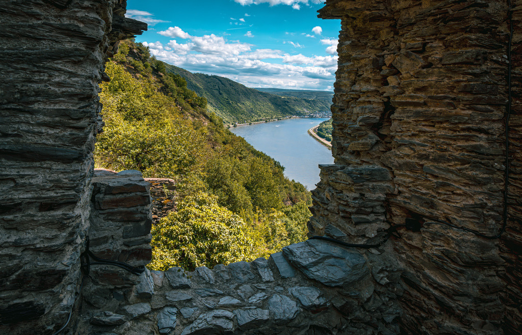 Blick durch Fenster der historischen Burg Liebenstein auf das Mittelrheintal | Der Blick aus dem Fenster der historischen Burg Liebenstein im UNESCO-Welterbe Oberes Mittelrheintal zeigt das malerische Rheintal bei Kamp-Bornhofen in Rheinland-Pfalz.Standort: Die Ruine liegt oberhalb des linken Rheinufers und bildet zusammen mit der nahegelegenen Burg Sterrenberg die als „Feindliche Brüder“ bekannten Burgengruppen.Aussicht: Durch die alten Schiefersteinmauern hindurch blickt man auf den bewaldeten Hang des Rheintals und den vorbeifließenden Fluss - Realisiert mit Pictrs.com