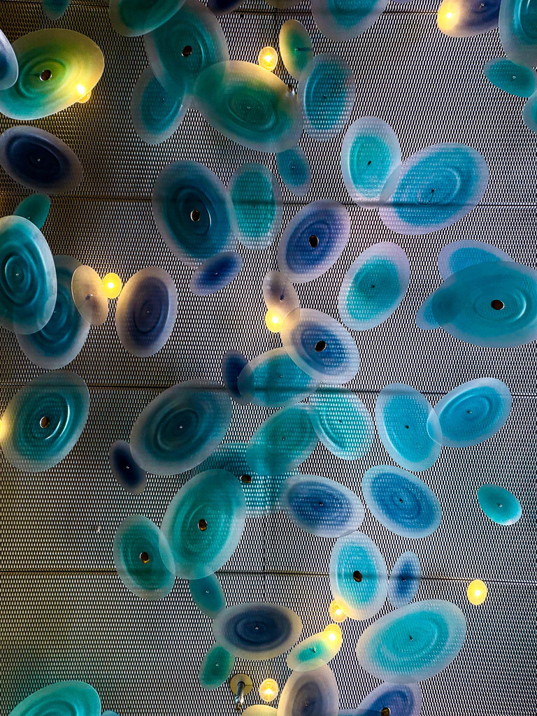 Neon-Biomorphismus: Abstraktes Lichtdesign im Club 3000 | Ein avantgardistisches Meisterwerk modernen Lichtdesigns. Diese faszinierende Architektur-Detailaufnahme fängt die lumineszierende Deckenleuchte des legendären Club 3000 in Nizza ein. Die organischen, zellartigen Formen in kühlem Neon-Blau und Aqua scheinen schwerelos vor einem geometrischen Raster zu schweben, während warme, goldene Lichtpunkte einen spannungsgeladenen Kontrast erzeugen. Dieses abstrakte Kunstwerk verbindet futuristischen Cyberpunk-Look mit biomorpher Eleganz. Ein absolutes High-End-Statement-Piece für moderne Wohnräume, Lounges oder kreative Büros. Perfekt zur Geltung gebracht als glänzender Acrylglas-Print für maximale Tiefenwirkung und Farbbrillanz. - Realisiert mit Pictrs.com