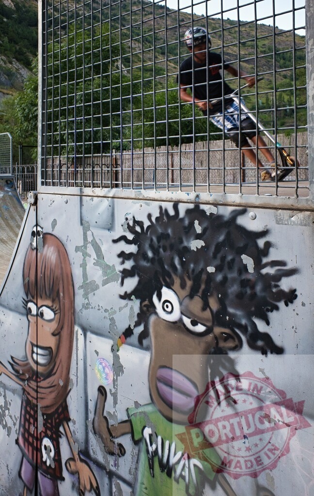 Scooter rider through fence at skatepark with graffiti art | Youth on a scooter riding a ramp behind a fence with vibrant street art characters on a wall below - Realized with Pictrs.com