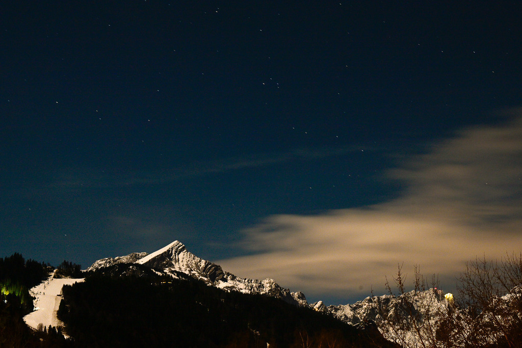 Nächtliche Wacht: Die Alpspitze unter dem Sternenzelt | Ein Bild von mystischer Erhabenheit und tiefer, winterlicher Stille: Inmitten der Dunkelheit thront der markante, schneebedeckte Gipfel der Alpspitze wie ein schweigender Wächter. Über dem schroffen Massiv spannt sich ein tiefblauer Nachtkristall-Himmel, auf dem unzählige Sterne wie feine Diamanten funkeln. Sanfte, von künstlichem Licht sanft illuminierte Wolkenbänder ziehen mystisch am Berghang vorbei, während links die hell erleuchtete Skipiste ins finstere Tal führt. Diese Aufnahme fängt die magische Poesie einer eisigen Winternacht in den Bergen ein – ein Moment voller Andacht, Einsamkeit und unendlicher Weite. - Realisiert mit Pictrs.com