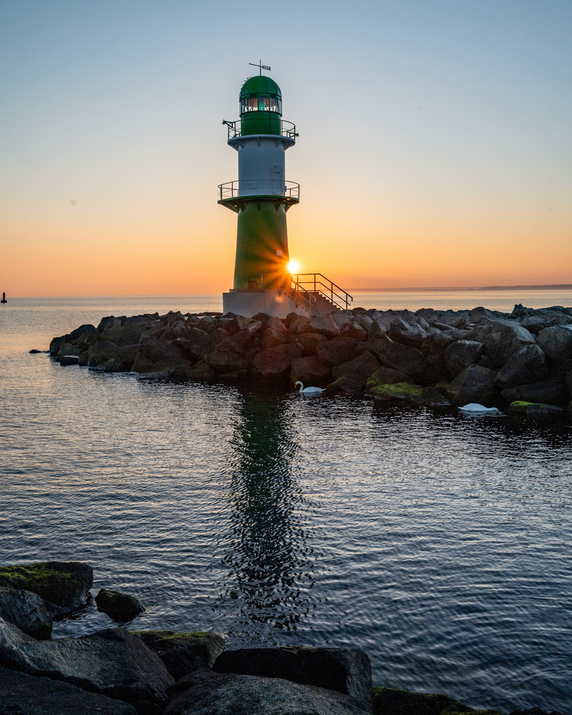 Westmolenfeuer Warnemünde im Morgendlicht | Das grüne Westmolenfeuer von Warnemünde steht im warmen Licht des Sonnenaufgangs auf der steinernen Mole. Der Sonnenstern am Fuß des Leuchtturms setzt einen markanten Lichtakzent. - Realisiert mit Pictrs.com
