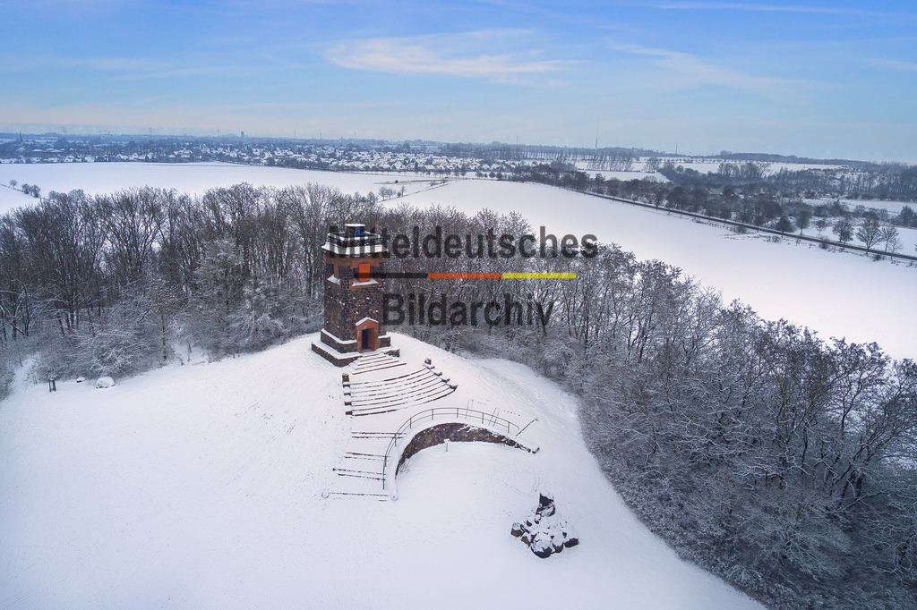 wartberg_niederndodeleben2_0018 | Dieses Luftbild zeigt den Bismarckturm auf dem Wartberg bei Niederndodeleben in Sachsen-Anhalt an einem klaren Wintermorgen. Der historische Aussichtsturm erhebt sich weithin sichtbar über die Landschaft der Magdeburger Börde und bildet das markante Hauptmotiv der Aufnahme. Aus der Vogelperspektive werden die besondere Lage des Bauwerks und die Weite der umliegenden Landschaft eindrucksvoll sichtbar.Die winterliche Stimmung verleiht dem Motiv eine außergewöhnliche Atmosphäre. Die Bäume rund um den Wartberg haben ihr Laub verloren und präsentieren sich als filigrane Strukturen in der frostigen Landschaft. Eine geschlossene Schneedecke überzieht Felder, Wege und Wiesen und verwandelt die Börde in eine ruhige, nahezu unberührte Winterlandschaft.Der klare Himmel und das kalte Morgenlicht sorgen für eine hervorragende Fernsicht und lassen die Konturen des Bismarckturms besonders deutlich hervortreten. Die Kombination aus schneebedeckter Landschaft, kahlen Bäumen und dem historischen Aussichtsturm verleiht der Aufnahme eine eindrucksvolle Ruhe und Klarheit.Dieses Luftbild zeigt den Bismarckturm von Niederndodeleben als markantes Wahrzeichen der Region und vermittelt einen stimmungsvollen Eindruck eines frostigen Wintermorgens in der Magdeburger Börde. - Realisiert mit Pictrs.com