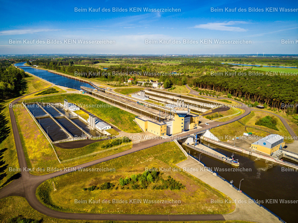 Doppelsparschleuse Hohenwarthe im Mittellandkanal-0010 | Das Luftbild zeigt die Doppelsparschleuse Hohenwarthe bei bestem Sommerwetter aus einer Drohnenperspektive. Die beiden parallel angeordneten Schleusenkammern sowie die markanten Sparbecken lassen die Dimensionen dieses bedeutenden Bauwerks des Wasserstraßenkreuzes Magdeburg eindrucksvoll erkennen. Aus der Vogelperspektive werden die klaren geometrischen Strukturen der Anlage und ihre Einbindung in den Elbe-Havel-Kanal besonders deutlich sichtbar.Die Doppelsparschleuse wurde 2003 in Betrieb genommen und überwindet einen Höhenunterschied von 18,55 bis 19,05 Metern zwischen dem Mittellandkanal und dem Elbe-Havel-Kanal. Jede der beiden Schleusenkammern besitzt eine Nutzlänge von 190 Metern und eine Breite von 12,50 Metern. Durch die seitlich angeordneten Sparbecken werden bei jedem Schleusenvorgang rund 60 Prozent des benötigten Wassers eingespart – ein wesentlicher Vorteil gegenüber konventionellen Schleusenanlagen. Die Drohnenaufnahme vermittelt einen eindrucksvollen Blick auf eines der modernsten Wasserbauwerke Deutschlands. Die Kombination aus leistungsfähiger Schleusentechnik, klarer Ingenieurarchitektur und der außergewöhnlichen Perspektive macht das Luftbild zu einer faszinierenden Dokumentation moderner Wasserstraßen- und Verkehrstechnik. - Realisiert mit Pictrs.com