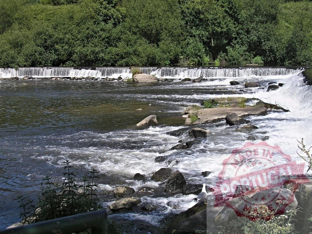 Water flowing over a weir creating an artificial waterfall. Norte - Portugal | River water churning and flowing over concrete and rocks, surrounded by green trees - Realized with Pictrs.com