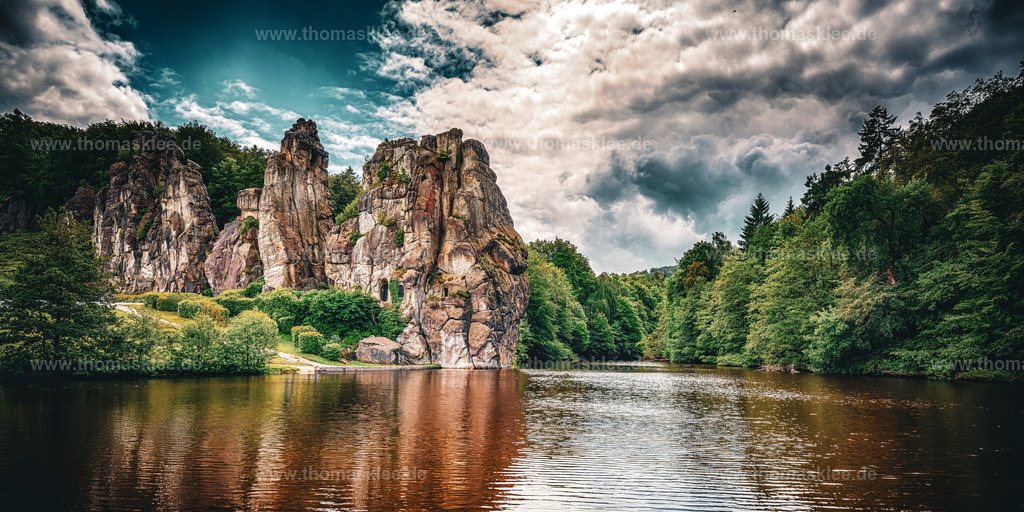 Mächtige Felsen im Teutoburger Wald Panorama | Dieses Panoramabild der Externsteine hat eine Wucht, die man sofort spürt. Das breite Format kommt dem Motiv perfekt entgegen — es gibt dem Blick Raum, sich zu entfalten, und lässt die mächtigen Sandsteinfelsen genau dort stehen, wo sie hingehören: im Mittelpunkt, ohne Gedränge, umgeben von Wald, Wasser und Himmel. Die Formation erhebt sich markant über dem dunklen See, ihre verwitterten Oberflächen in warmen Braun- und Ockertönen leuchtend vor dem dramatisch bewölkten Himmel. Was dieses Bild so besonders macht, ist das Zusammenspiel der Stimmungen. Der Himmel ist nicht einfach blau — er ist ein echtes Schauspiel aus schweren Wolken, die von warmem Licht durchbrochen werden. Diese Spannung zwischen Licht und Dunkel zieht sich durch das gesamte Bild. Das Wasser im Vordergrund nimmt die Farben des Himmels und der Felsen auf und spiegelt sie in einem tiefen, satten Rotbraun wider. Die Bäume rechts und links rahmen die Szene ein, ohne sie einzuengen. Die Externsteine im Teutoburger Wald gehören zu den bekanntesten Naturdenkmälern Deutschlands. Diese Sandsteinformation in Nordrhein-Westfalen war über Jahrtausende ein Ort von spiritueller und kultureller Bedeutung — von vorchristlichen Kultstätten bis hin zu frühmittelalterlichen Felsreliefs. Diese Geschichte spürt man in einem Bild wie diesem, auch ohne sie zu kennen. Es liegt etwas Zeitloses in der Art, wie die Felsen dort stehen — als hätten sie schon immer da gestanden und als würden sie noch lange da stehen .Fotografisch ist das Bild mit einem kräftigen, modernen Stil bearbeitet: gesättigte Farben, starker Kontrast, eine leicht cinematische Anmutung. Das macht es zum idealen Wandbild für große Formate — ein Bild, das einen Raum füllt und eine Geschichte erzählt. - Realisiert mit Pictrs.com