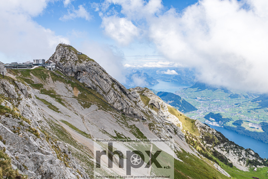 Pilatus Gipfelgrat mit Vierwaldstättersee, Zentralschweiz | Blick entlang des felsigen Gipfelgrats des Pilatus (2132 m ü. M.) auf den Hotelkomplex Pilatus-Kulm. Im Hintergrund der Vierwaldstättersee mit umliegenden Ortschaften, grünen Hügeln und Alpenpanorama. Sommerbedingungen mit Quellwolken. Kanton Nidwalden/Obwalden/Luzern, Schweiz. - Realisiert mit Pictrs.com