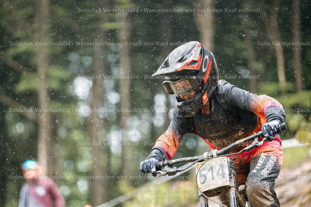 Mountainbiker im Regen | Ein Mountainbiker in voller Montur rast einen staubigen Trail hinunter. Er trägt einen roten Helm mit schwarzen Akzenten, eine dunkle Brille und einen schwarzen Trikot mit dem Schriftzug 'TREK'. Auf seinem Rücken befindet sich eine Nummer 514. Es regnet leicht, und der Trail ist mit Staub bedeckt. Im Hintergrund sind Bäume und Büsche zu sehen. - Realisiert mit Pictrs.com