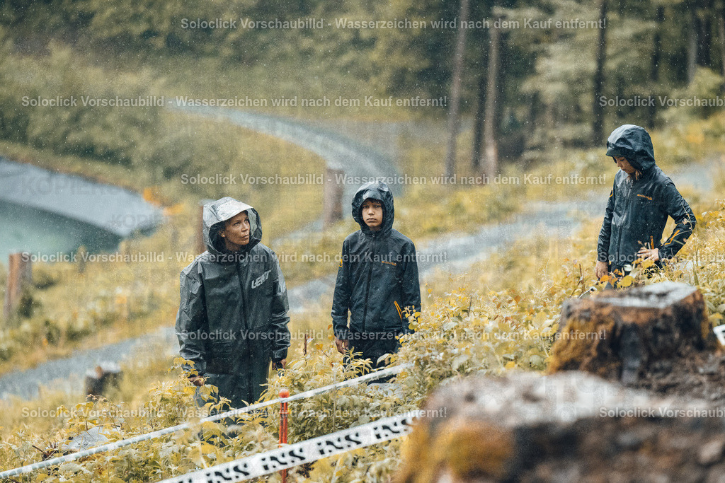 Regenwanderung im Wald | Das Foto zeigt drei Personen, vermutlich Wanderer oder Naturforscher, die im Regen stehen. Sie tragen alle wasserdichte Jacken und Hosen und schauen in Richtung einer schmalen, kurvenreichen Straße, die durch einen dichten Wald führt. Das Gelände ist mit hohem Gras und zahlreichen Baumstümpfen bedeckt. Ein gelbes Absperrband ist im Vordergrund zu sehen. - Realisiert mit Pictrs.com