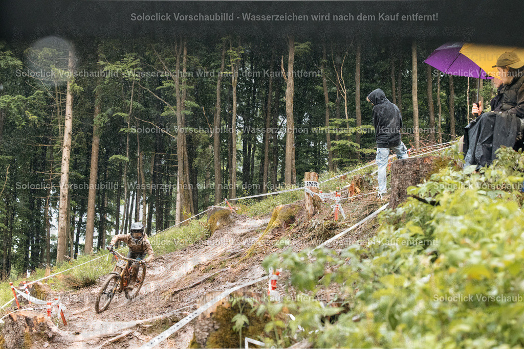 Mountainbike-Abenteuer im Wald | Das Bild zeigt einen Mountainbiker, der einen anspruchsvollen, kurvigen Pfad in einem dichten Wald hinabfährt. Der Pfad ist steil und mit Wurzeln und Steinen übersät. Der Fahrer trägt einen Helm und Schutzkleidung. Am Rand des Pfades steht ein Zuschauer mit einem Regenschirm, der das Rennen beobachtet. Die Szene vermittelt ein Gefühl von Abenteuer und Herausforderung. - Realisiert mit Pictrs.com