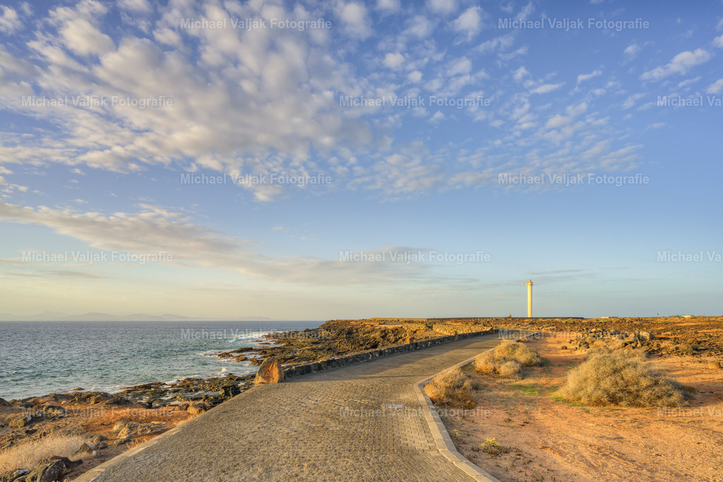 Weg zum Leuchtturm am Punta Pechiguera auf Lanzarote | Ein gepflasterter Weg zieht sich elegant an der Küste entlang, während links das Meer in gleichmäßigen Wellen gegen die Felsen schlägt. Die klare Linie des Weges wirkt wie eine ruhige Führung durch diese karge Umgebung. Rechts breitet sich das trockene, helle Vulkangelände aus, links öffnet sich die Weite des Atlantiks. Am Ende der Perspektive steht der Leuchtturm von Pechiguera – schlank, hell und unübersehbar –, als würde er den Blick des Betrachters bewusst zu sich hinziehen. - Realisiert mit Pictrs.com