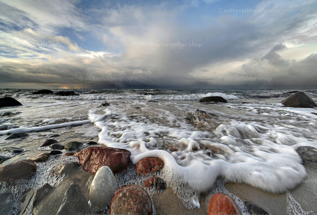 Schaumkronen vor aufziehender Regenfront | Schaumkronen umspülen die bunten, runden Steine am Strandsaum, während am Horizont eine Wolke bedrohlich näherkommt.
