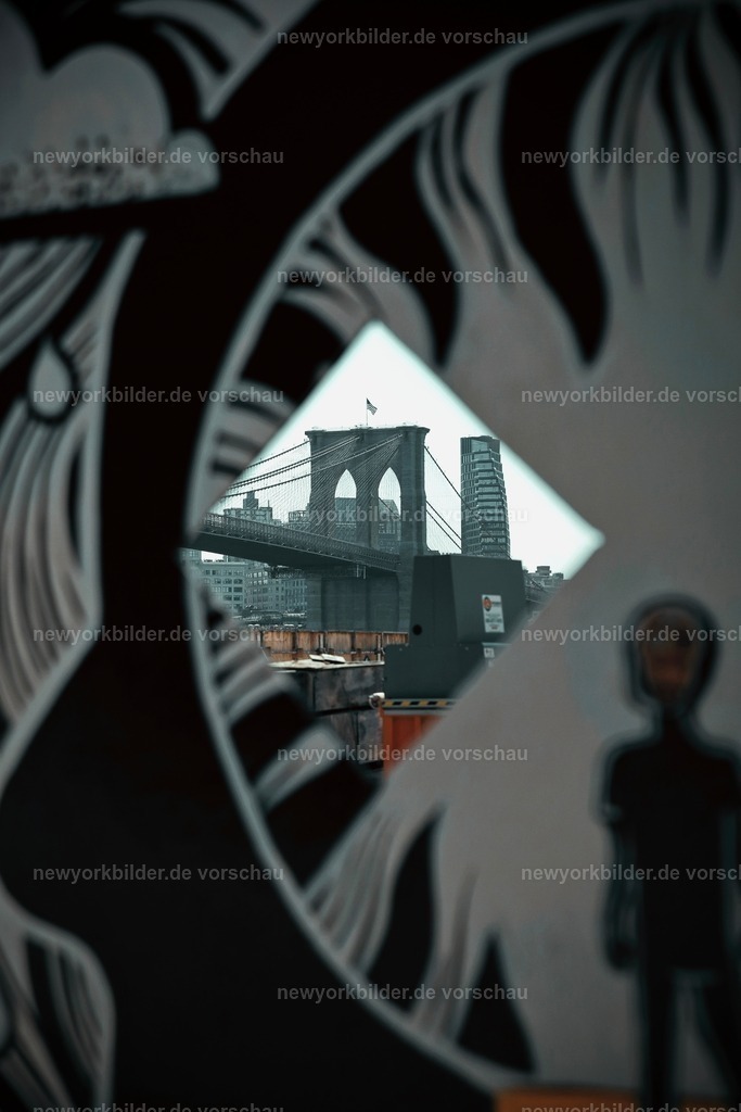 Magischer Blick auf die Brooklyn Bridge | Die Brooklyn Bridge ist mehr als nur eine Brücke – sie ist ein Symbol für Freiheit, Geschichte und die pulsierende Energie New Yorks. Doch manchmal sind es die versteckten Perspektiven, die den Zauber ausmachen. Am East River verbirgt ein Holzbauzaun den freien Blick, aber ein kleines Fensterloch eröffnet eine magische Sicht auf die Brooklyn-Seite der Brücke. Durch diesen geheimen Rahmen entsteht ein Foto, das die ikonische Architektur in einem völlig neuen Licht zeigt  – ein Augenblick, der bleibt.