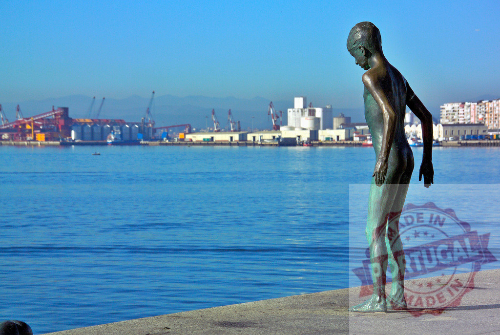Bronze boy statue looking at industrial port panorama. Spain - Cantabria | Bronze statue of a boy standing on a pier, observing an industrial seaport across the water - Realisiert mit Pictrs.com
