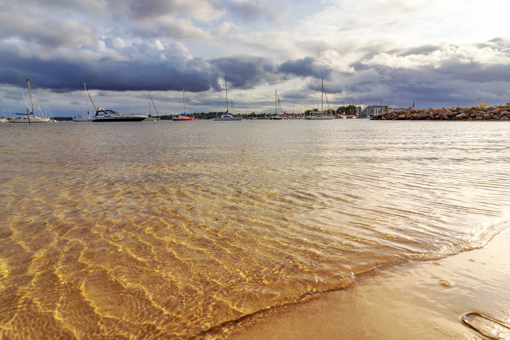 Wandbild: Strand in Eckernförde | Dieses Wandbild im Querformat zeigt den Strand in Eckernförde. Im Vordergrund ist das flache Wasser direkt am Strand zu sehen. Durch die abendliche Sonne entstehen schöne Reflexionen im Wasser. Am Himmel sind viele dunkle Wolken zu sehen.  - Realisiert mit Pictrs.com