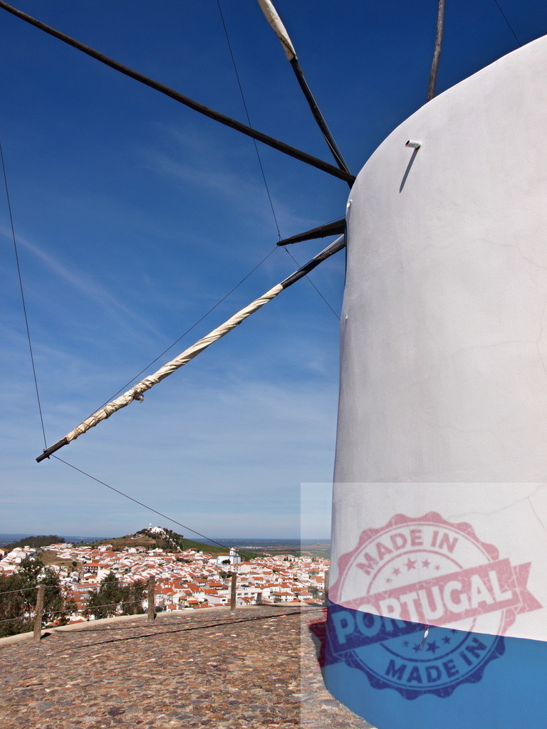 Traditional white windmill on hilltop overlooking Alentejo village | Old windmill standing on a cobbled path overlooking a traditional Portuguese village under clear blue sky - Realized with Pictrs.com