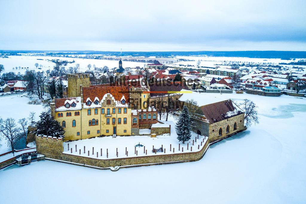 Schloss Flechtingen mit See im Winter-0020 | Dieses Luftbild zeigt das Wasserschloss Flechtingen im Landkreis Börde in Sachsen-Anhalt während eines winterlichen Tages. Die historische Schlossanlage präsentiert sich in einer ruhigen und nahezu märchenhaften Atmosphäre. Eine geschlossene Schneedecke bedeckt die Dächer, Wege und Parkflächen, während auch der umliegende Schlosssee von Eis und Schnee überzogen ist.Das Wasserschloss Flechtingen zählt zu den bekanntesten Schlossanlagen Sachsen-Anhalts und prägt mit seiner Lage am See das Ortsbild des Luftkurortes Flechtingen. Die Kombination aus historischer Architektur, Wasserlandschaft und weitläufigen Parkanlagen macht das Schloss zu einem beliebten Ausflugsziel und einem bedeutenden Kulturdenkmal der Region.Aus der Vogelperspektive werden die harmonischen Strukturen der Anlage besonders deutlich sichtbar. Der gefrorene Schlosssee umrahmt das Gebäude und verleiht der Aufnahme eine außergewöhnliche Wirkung. Die winterliche Landschaft betont die historischen Formen des Schlosses und schafft einen reizvollen Kontrast zwischen Natur und Architektur.Dieses Luftbild zeigt das Wasserschloss Flechtingen in einer besonderen Jahreszeit und vermittelt einen eindrucksvollen Eindruck von der Schönheit historischer Bauwerke und Winterlandschaften in Sachsen-Anhalt. - Realisiert mit Pictrs.com