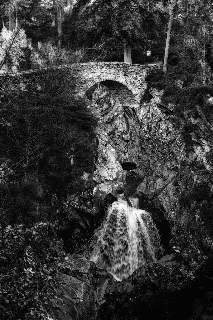Historische Steinbrücke, Falls of Bruar | Eine historische Steinbogenbrücke überspannt einen felsigen Wasserfall, umgeben von dichtem Wald. Die Schwarz-weiss-Darstellung betont die Texturen des Gesteins und die fliessende Bewegung des Wassers, was eine zeitlose und dramatische Ästhetik schafft. Die vertikale Komposition unterstreicht die Höhe des Wasserfalls und die Integration der Brücke in die natürliche Landschaft. - Realisiert mit Pictrs.com