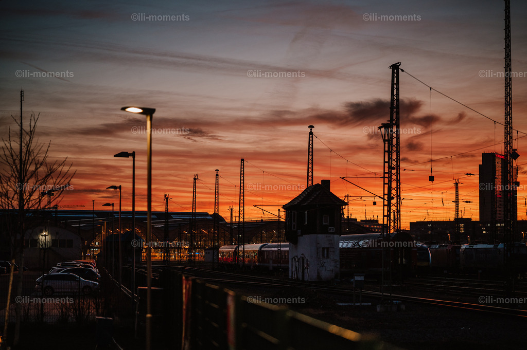 Kommen & Gehen | „Kommen &amp; Gehen“ ist ein eindrucksvolles urbanes Fotokunstwerk, das die besondere Stimmung eines Großbahnhofs in der Abenddämmerung einfängt. Das Bild vereint zwei Ebenen des Abschieds und Neubeginns: das Kommen und Gehen der untergehenden Sonne, deren warmes Abendlicht die Szenerie in weiche, gold-rote Töne taucht – und das Kommen &amp; Gehen der Menschen, das man unweigerlich mit dem pulsierenden Bahnhofsgeschehen verbindet.Die moderne Architektur, weitläufigen Bahnsteige und die sichtbaren Gleisstrukturen vermitteln Bewegung und Dynamik, während das Abendlicht zugleich Ruhe und Tiefe ausstrahlt. Diese Kombination aus Urbanität und Stimmung macht das Foto zu einem idealen Kunstwerk für moderne Wohnräume, Büros, Warte- und Empfangsbereiche oder überall dort, wo man dem Betrachter ein Gefühl von Reise, Übergang und Zeit vermitteln möchte. - Realisiert mit Pictrs.com