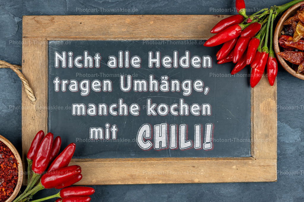 Chili-Spruch auf Schiefertafel - Küchenweisheit | Eine rustikale Schiefertafel in einem Holzrahmen zeigt einen witzigen Spruch über Chili-Liebhaber. Frische rote Chilischoten schmücken die Ecken der Tafel und verleihen dem Arrangement lebendige Farbtupfer. Zwei Holzschalen mit getrockneten Chilis und Gewürzpulver ergänzen die Komposition perfekt. Der humorvolle Text "Nicht alle Helden tragen Umhänge, manche kochen mit CHILI!" bringt Küchenfreunde zum Schmunzeln. Das Bild strahlt Gemütlichkeit und Leidenschaft für scharfe Küche aus. - Realisiert mit Pictrs.com