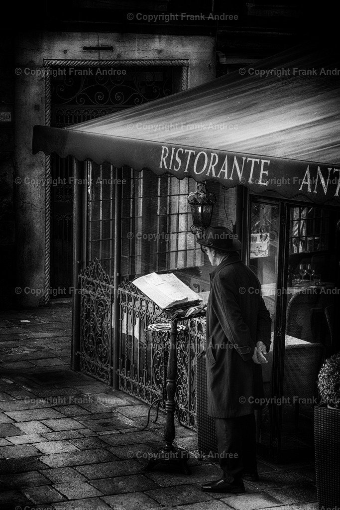 Venedig | Ein Mann mit Hut und Mantel steht vor einem Restaurant in Venedig. Straßenfotografie aus Venedig in schwarz weiß. - Realisiert mit Pictrs.com