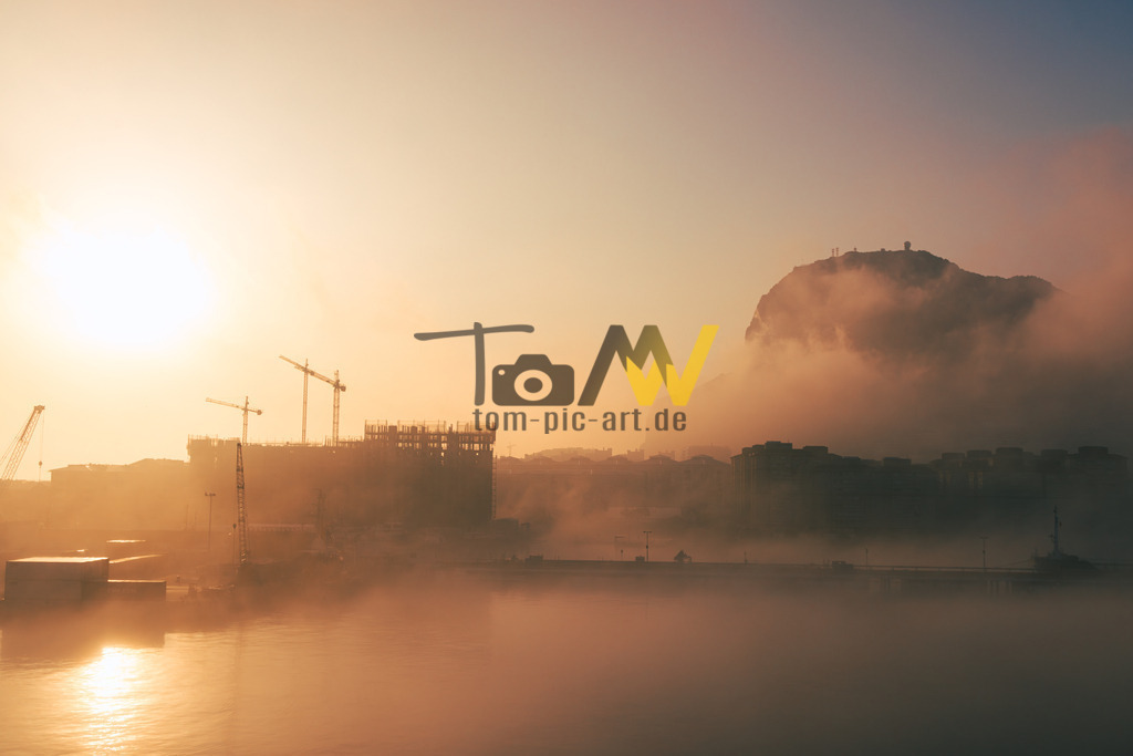 Mystischer Sonnenaufgang im Hafen von Gibraltar-Dunst und Nebel | Die Aufnahme zeigt eine neblige Morgendämmerung über einem Gewässer mit einer Baustelle und Kränen im Hintergrund.Die Sonne scheint hell am Horizont und erzeugt ein warmes, goldenes Licht.Dichter Nebel oder Dunst liegt über der Wasseroberfläche und den unteren Bereichen der Landschaft. - Realisiert mit Pictrs.com