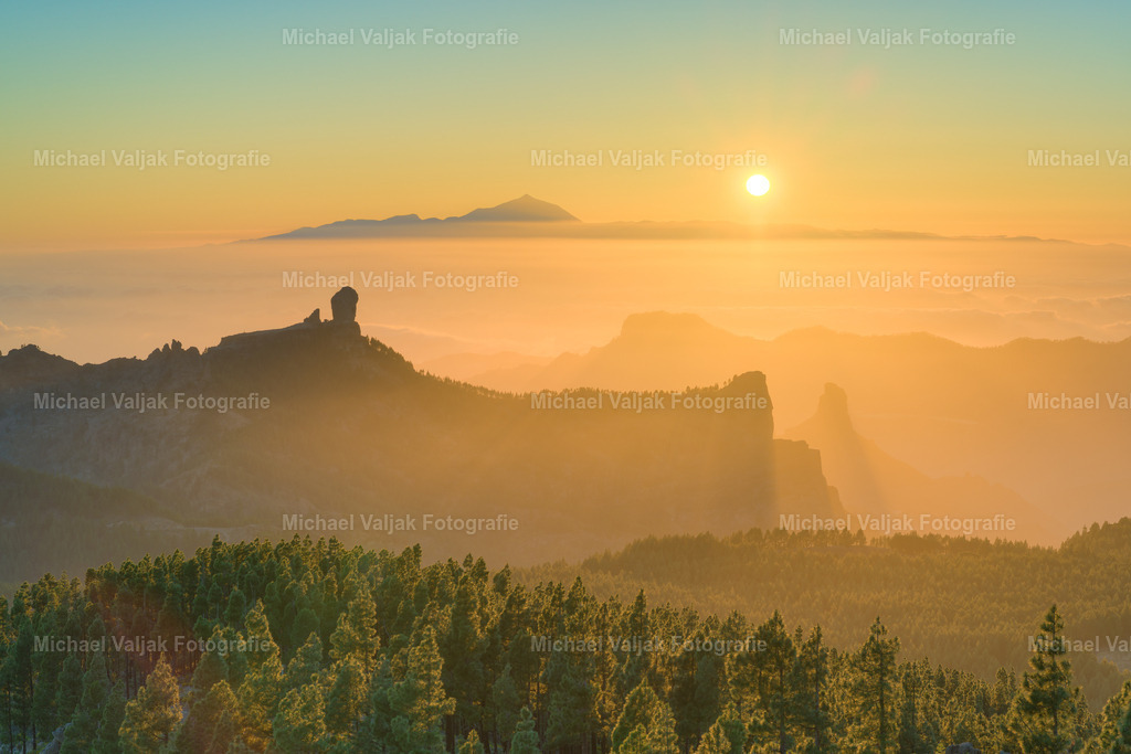 Mirador del Pico de Las Nieves Gran Canaria | Vom Pico de las Nieves aus eröffnet sich bei Sonnenuntergang ein weiter Blick über die Bergwelt Gran Canarias. Die zerklüfteten Landschaften liegen im warmen Licht der tief stehenden Sonne, während sich Schatten in den Tälern ausbreiten. In der Ferne zeichnen sich markante Felsformationen wie der Roque Nublo ab. Am Horizont erhebt sich Teneriffa mit dem Teide aus einem dichten Wolkenmeer – fast wie eine Insel inmitten des Himmels. Der Himmel leuchtet in sanften Orange- und Rottönen und lässt die raue Vulkanlandschaft in einem weichen Licht erscheinen – ein stiller Moment über den Wolken. - Realisiert mit Pictrs.com