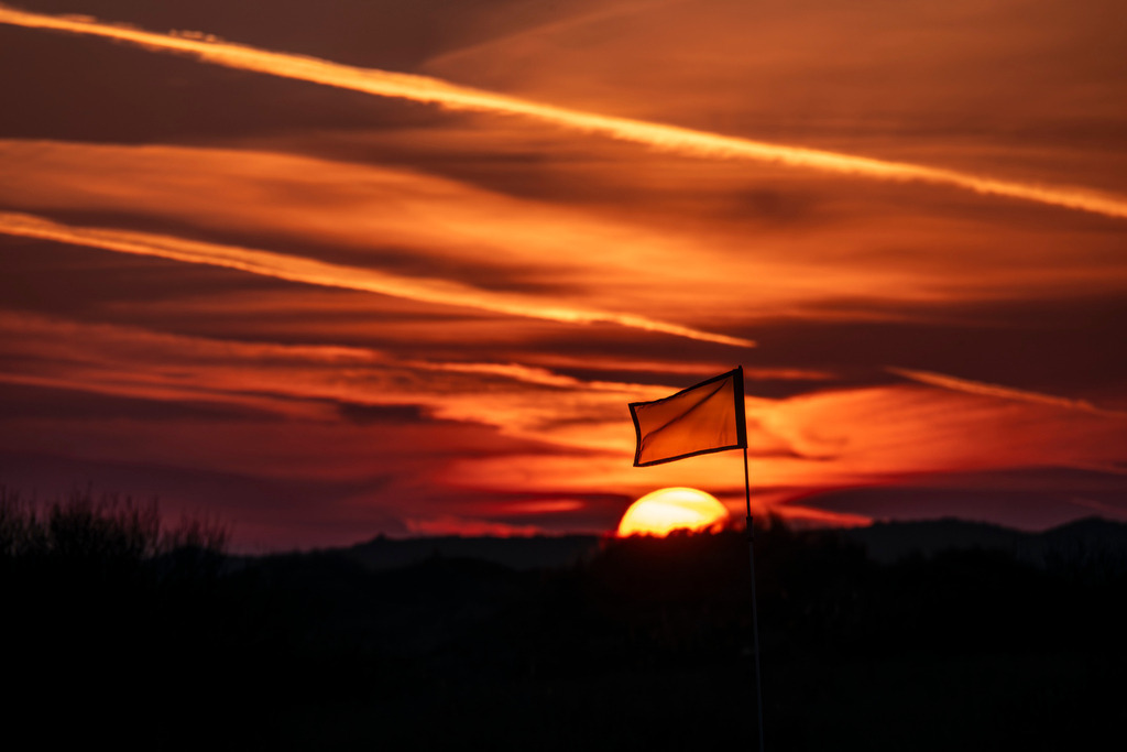 Sonnenuntergang auf Golfplatz | Eine stimmungsvolle Aufnahme eines intensiven Sonnenuntergangs auf der Insel Texel. Im Vordergrund steht die Silhouette einer kleinen Fahne an einem Mast, die vor der untergehenden Sonne positioniert ist. Der Himmel ist in dramatische Rot- und Orangetöne getaucht, während markante Kondensstreifen von Flugzeugen die Wolkenstruktur durchbrechen und dem weiten Horizont eine grafische Tiefe verleihen. - Realisiert mit Pictrs.com