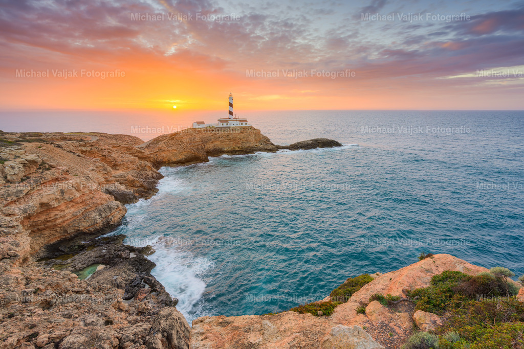Sonnenaufgang am Faro de Cala Figuera auf Mallorca | Am Faro de Cala Figuera öffnet sich der Blick über eine kleine Bucht hinweg in Richtung Leuchtturm und Sonnenaufgang. Der Himmel leuchtet in satten Orange‑ und Gelbtönen, die sich klar über der offenen Küstenlinie abzeichnen. Das warme Morgenlicht legt sich gleichmäßig auf die Felsen, während das Meer in der Bucht ruhig bleibt und erste Reflexe der kräftigen Farben aufnimmt. Die Kombination aus weiter Sicht, klarer Lichtstimmung und der markanten Küstenform erzeugt einen sachlichen, präzisen Eindruck dieses frühen Moments. - Realisiert mit Pictrs.com