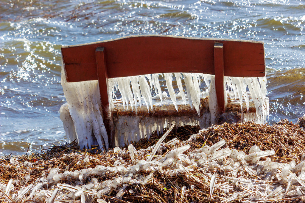 Wandbild: Vereiste Bank in Lindaunis an der Schlei | Dieses Wandbild im Querformat zeigt eine vereiste Bank im Uferbereich der Schlei in Lindaunis. Direkt hinter der Bank sind die Wellen der Schlei in schöner Lichtstimmung zu sehen.  - Realisiert mit Pictrs.com