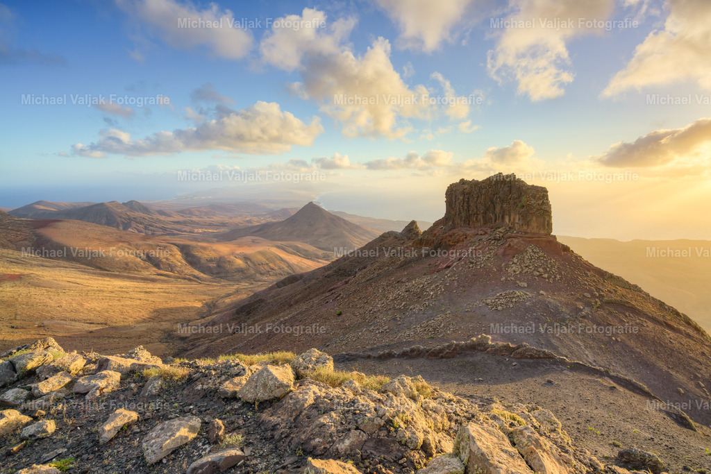 Blick vom Montana Cardon auf Fuerteventura | Vom Montaña Cardón reicht der Blick weit über die karge Landschaft des Südens. Die Hänge fallen sanft ab, die Ebenen darunter wirken offen und weit. In der Ferne ziehen sich die Berge wie eine ruhige Linie durch die Insel. Ein stiller Moment, der zeigt, wie viel Raum und Klarheit Fuerteventura von oben ausstrahlt. - Realisiert mit Pictrs.com