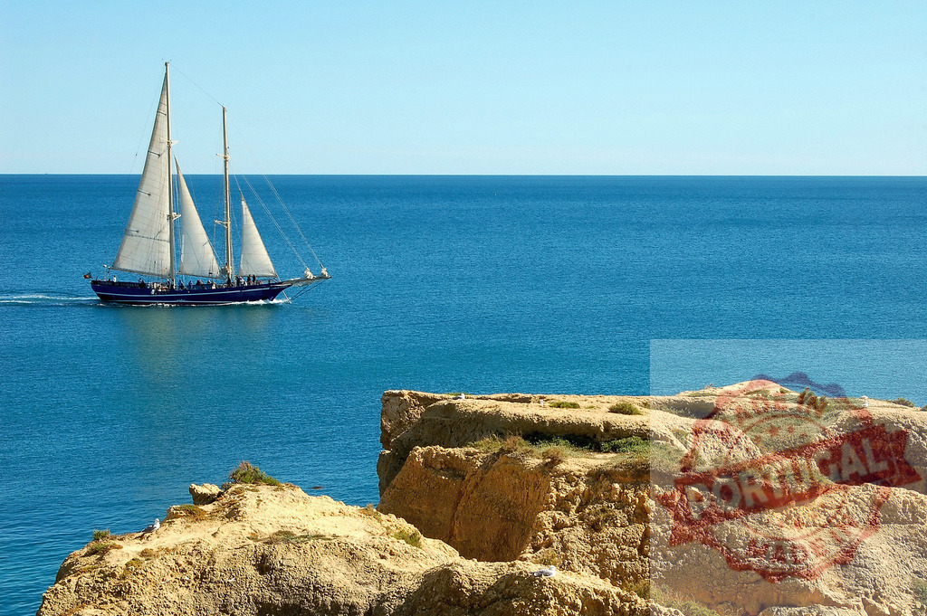Sailboat cruising blue ocean near rugged cliffs | Sailboat cruising along a calm blue ocean next to a textured coastline with cliffs - Realized with Pictrs.com