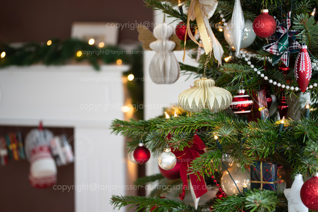 Weihnachtsbaum Wabenschmuck Wohnzimmer D | Geschmückter Weihnachtsbaum, echte Nordmanntanne vor unscharfem Wohnzimmermöbel. Makrofotografie zeigt Weihnachtsbaum Ornamente aus Waben Papier und Kugeln in Rot, Weiß und Champagner.