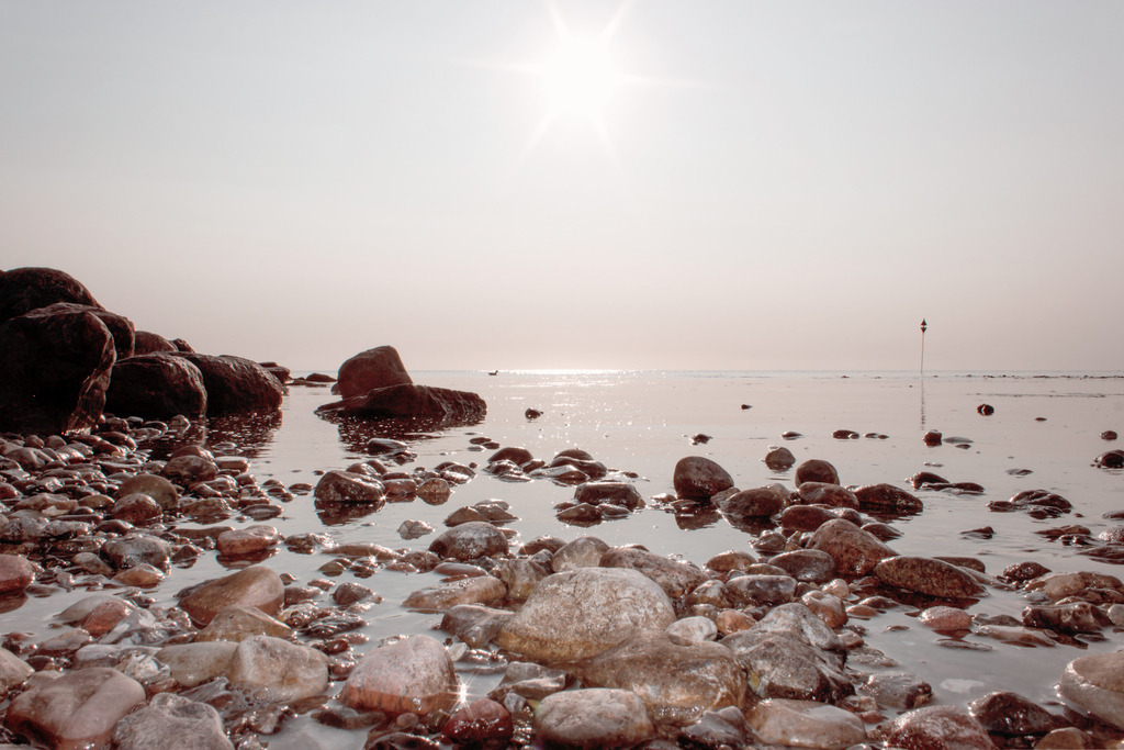 Wandbild: Sonnenschein über der Ostsee | Dieses Wandbild im Querformat zeigt die Sonne mit schönen Sonnenstrahlen über dem Meer. Der Himmel wirkt durch den Sonnenschein beige. Im Vordergrund liegen einige Steine im Meer. Auf der linken Seite sind einige dunklere Steine der Steinmole zu sehen. Holen Sie sich dieses traumhafte Strandmotiv in dezenten Farben auf Leinwand, auf Aluminium-Platte oder Acrylglas. Ideal fürs Wohnzimmer, Schlafzimmer, Küche, den Arbeitsplatz oder die Ferienwohnung. Die Wandbilder werden individuell für Sie in vielen Abmessungen produziert. Daher passen die Ostseekult Wandbilder immer perfekt an Ihre Wände.  - Realisiert mit Pictrs.com