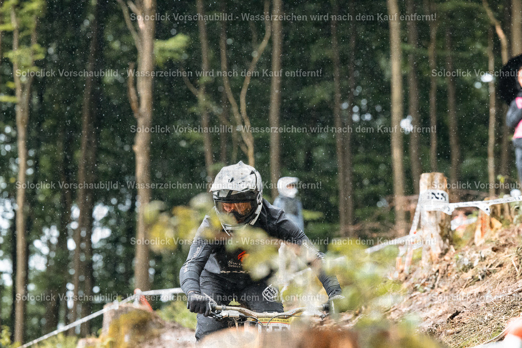 Adrenalin im Wald | Ein Mountainbiker in voller Ausrüstung rast mit hoher Geschwindigkeit einen steilen, kurvigen Pfad in einem dichten Wald hinunter. Der Fahrer trägt einen schwarzen Helm mit einer dunklen Visier und eine schwarze Jacke mit dem Schriftzug 'Swiss'. Er greift die Bremsen und lenkt geschickt, um die Kontrolle über das Bike zu behalten. Der Hintergrund besteht aus dichtem Grün und braunen Baumstämmen, was die Wildnis des Geländes unterstreicht. Es regnet leicht, was die Szene noch dramatischer macht. - Realisiert mit Pictrs.com