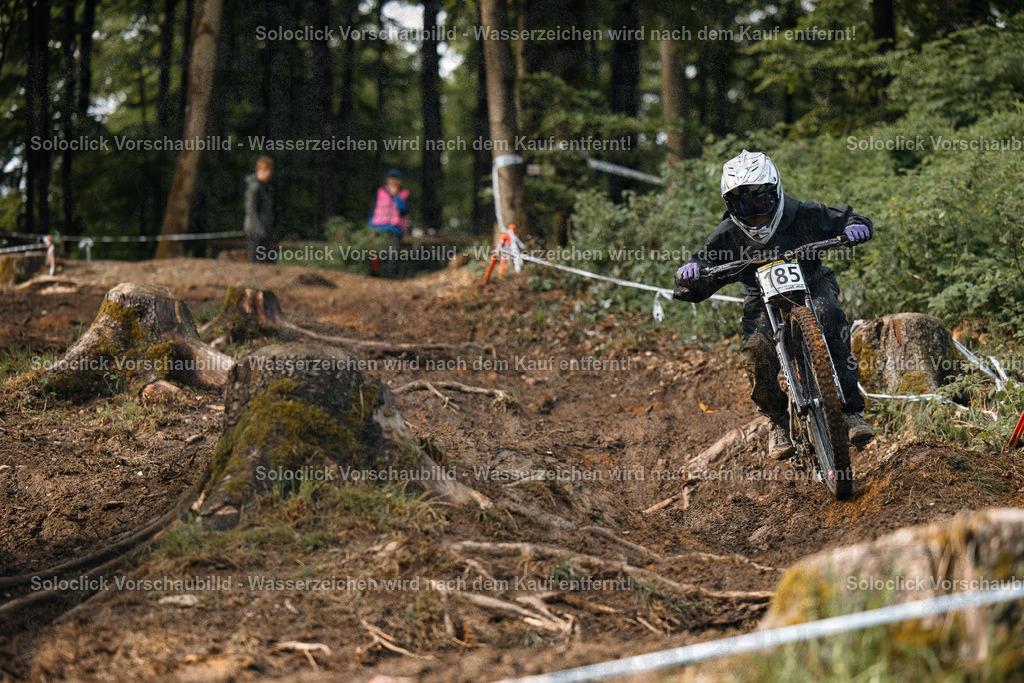 Downhill-Action im Wald | Das Bild zeigt einen Mountainbiker in voller Montur, der einen anspruchsvollen Trail in einem bewaldeten Gebiet durchquert. Der Biker trägt einen schwarzen Helm mit einer weißen Markierung und eine dunkle Weste mit dem Nummernschild 85. Er fährt einen Downhill-Mountainbike mit dicker Federung und breiten Reifen. Der Trail ist steil, mit vielen Wurzeln, Steinen und Kurven. Der Wald ist dicht und die Bäume sind hoch. Im Hintergrund sind weitere Personen zu sehen, die den Biker beobachten. - Realisiert mit Pictrs.com
