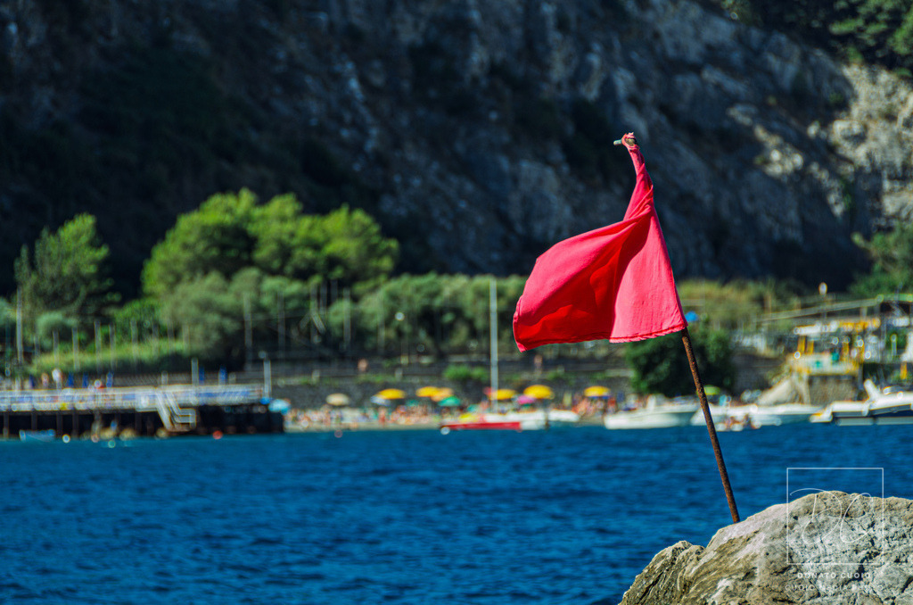 Amalfi Red | Ein rotes Signal vor mediterraner Postkartenkulisse – eingefangen in einer malerischen Bucht von Amalfi. Diese Fotografie vereint Symbolkraft mit mediterranem Charme: Die rote Flagge steht für Wachsamkeit, Respekt und Naturgewalt – mitten im Sommerparadies.Ein Fine-Art-Motiv für moderne Räume, das nicht nur schön aussieht, sondern auch Fragen stellt. - Powered by Pictrs.com