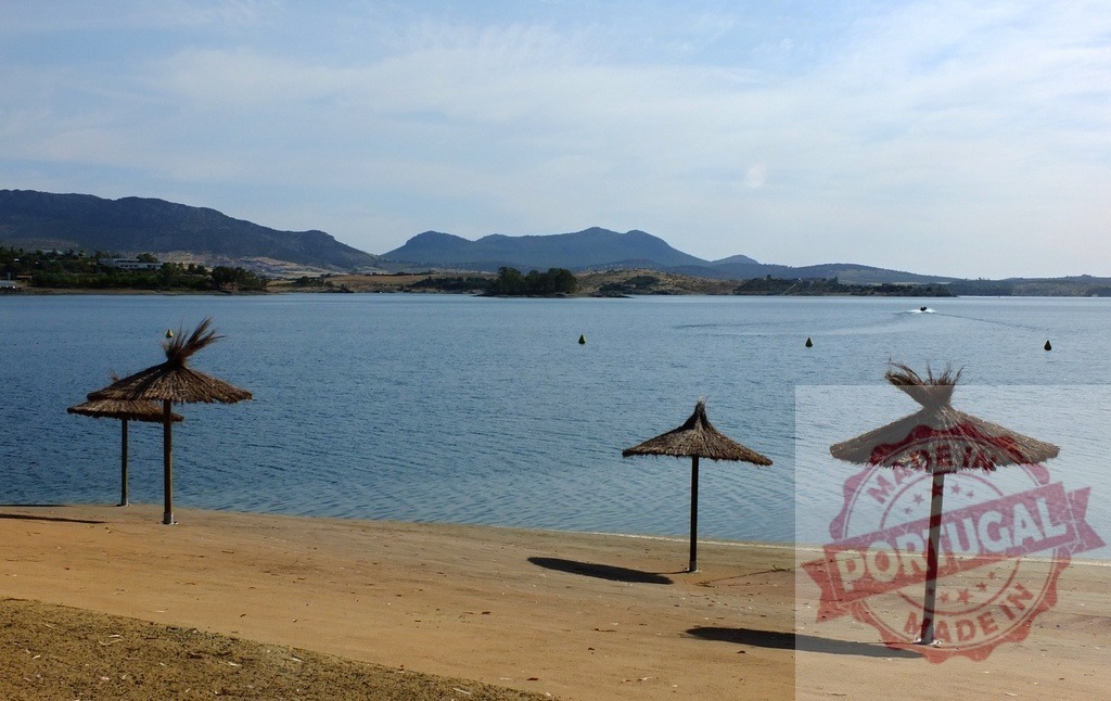 Straw beach umbrellas on lake shore with mountains. Spain - Extremadura | Straw umbrellas providing shade on a peaceful lake beach with distant mountains - Realized with Pictrs.com