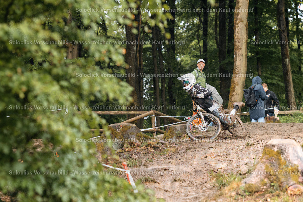 Mountainbike-Action im Wald | Ein Mountainbiker rast auf einem schlammigen Pfad durch einen dichten Wald. Er trägt einen Helm und Schutzkleidung und scheint sich in vollem Tempo zu bewegen. Im Hintergrund sind weitere Personen zu sehen, die den Fahrer beobachten. Die Szene vermittelt ein Gefühl von Abenteuer und Action. - Realisiert mit Pictrs.com