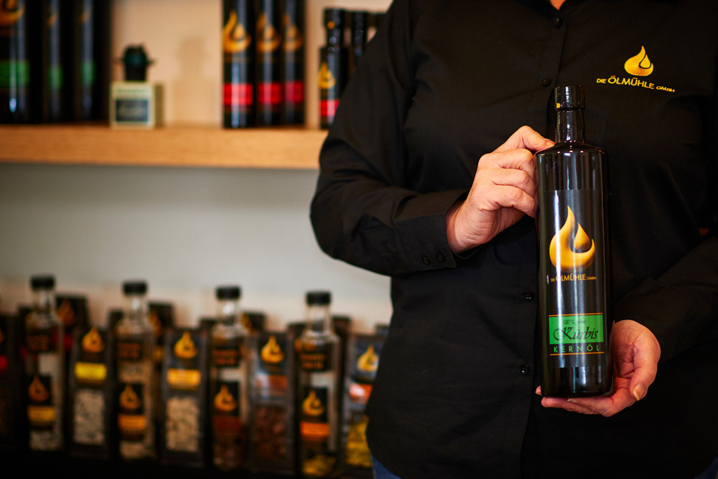 Die Ölmühle | Markt Allhau, Austria - February 09, 2018: Die Oelmuehle; Geschäftsführerin Helga Gradwohl präsentiert eine Flasche Kernöl vor dem Verkaufsregal. - Realisiert mit Pictrs.com