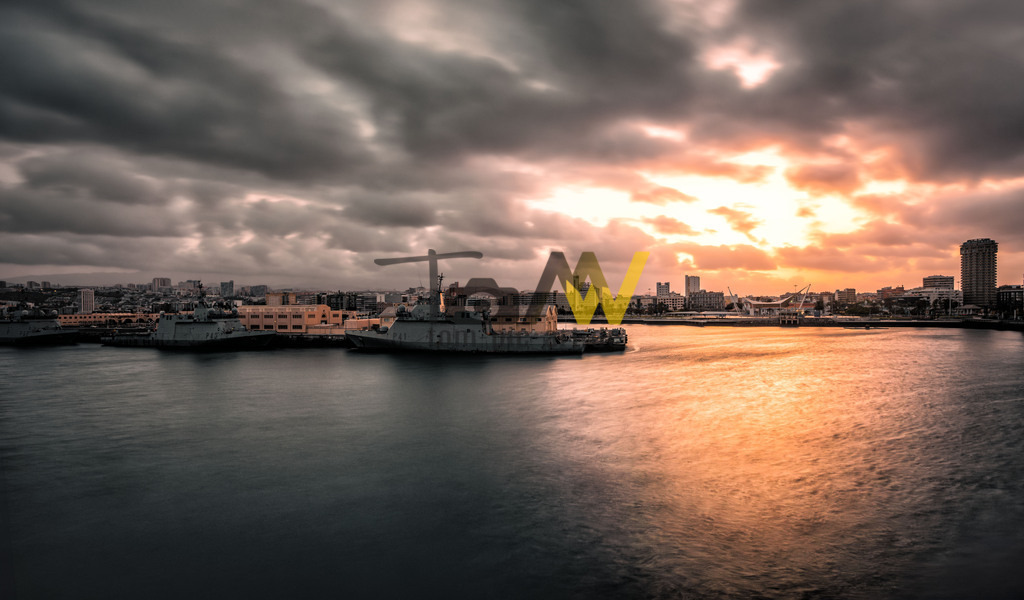 Sonnenuntergang in Las Palmas de Gran Ganaria,Puerto de La Luz | Das Bild zeigt den Hafen von Las Palmas de Gran Canaria bei Sonnenuntergang. Las Palmas de Gran Canaria ist die Hauptstadt der gleichnamigen Insel und die größte Stadt der Kanarischen Inseln. Die Stadt ist durch einen der größten Häfen Spaniens geprägt, den "Puerto de La Luz". Der Hafen befindet sich auf der Nordostseite der Insel, wo eine kleine Halbinsel ins Meer ragt. Das Bild fängt einen dramatischen Moment mit stark bewölktem Himmel und der goldenen Stunde des Sonnenuntergangs über der Skyline der Stadt ein. - Realisiert mit Pictrs.com