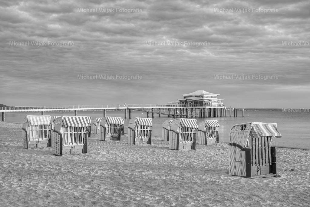 Morgensonne am Timmendorfer Strand schwarz-weiß | An einem bewölkten Morgen schafft es die Sonne kurz durch die Wolkendecke und taucht die Seeschlösschenbrücke mit dem Teehaus am Ende und die Strandkörbe am Strand in ein warmes Licht (Schwarz-weiß-Version).  - Realisiert mit Pictrs.com