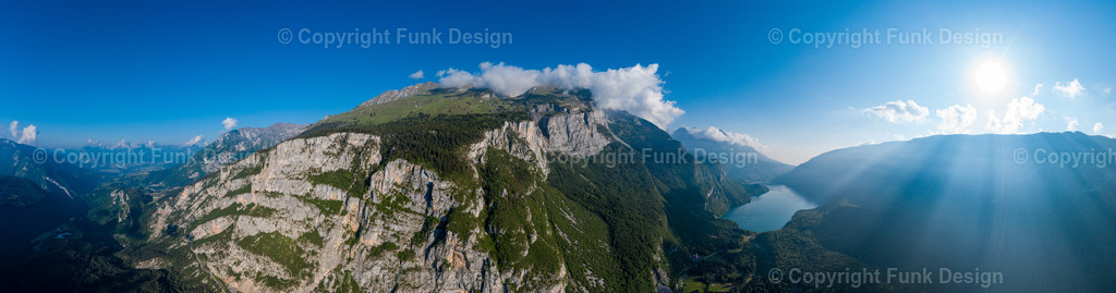 Sonnenstrahlen über den Bergen – Trentino, Italien | Ein weites Drohnenpanorama zeigt eine mächtige Felswand und dahinter ein Tal mit See, während die Sonne rechts im Bild starke Lichtstrahlen über die Berge wirft. Die Szene wirkt episch, frei und klar und eignet sich perfekt als großes Panorama-Wandbild.